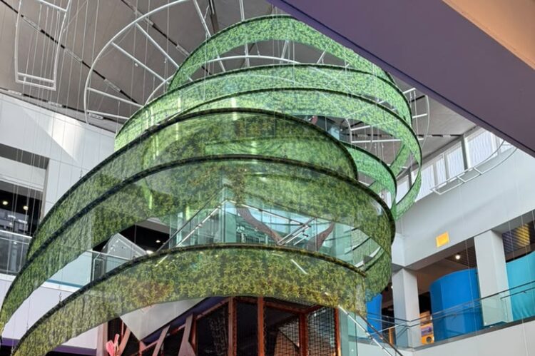 Indoor atrium with a vertical green spiral mesh sculpture over a wooden coral-like exhibit and information panels.